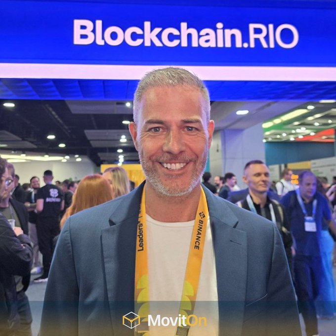 A man wearing a blue blazer and a Binance lanyard smiles at the Blockchain.RIO conference, with a MovitOn watermark at the bottom.