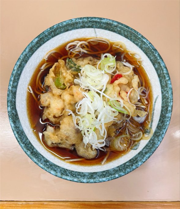 A bowl of soba noodles in broth topped with tempura, shredded scallions, and a bit of red chili, served in a blue-rimmed ceramic bowl on a peach-colored surface.