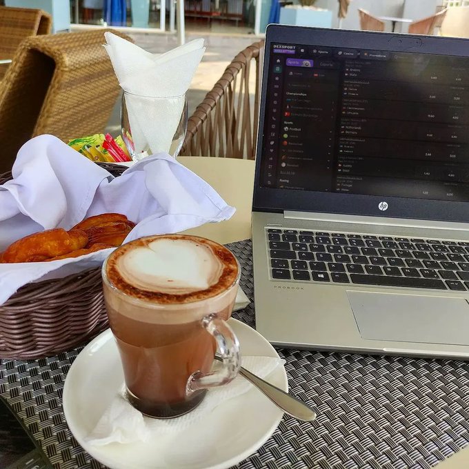 A coffee cup with a heart design in the foam sits next to an HP ProBook laptop displaying a cryptocurrency trading interface, with a basket of bread nearby.