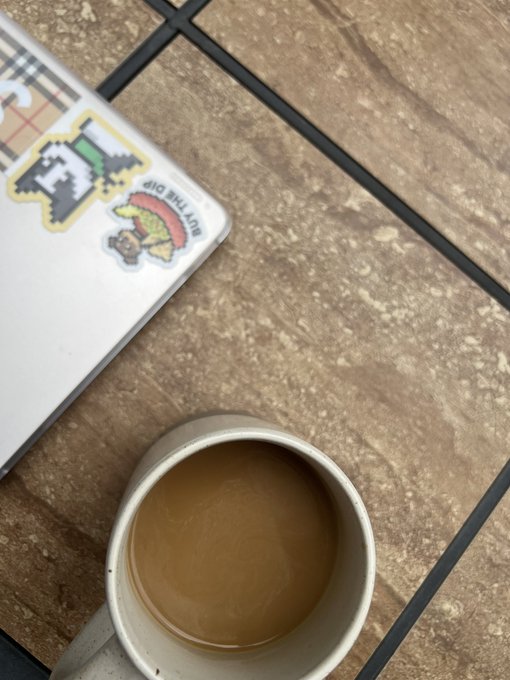 A top-down view of a mug of coffee with cream next to a laptop covered in stickers, including one that says "Buy This Dip" with a pixel art Bitcoin character.