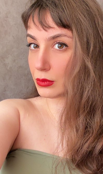 A close-up selfie portrait of a young woman with long light brown hair, wispy bangs, striking eyes, and bright red lipstick, wearing an olive green strapless top against a textured gray wall.