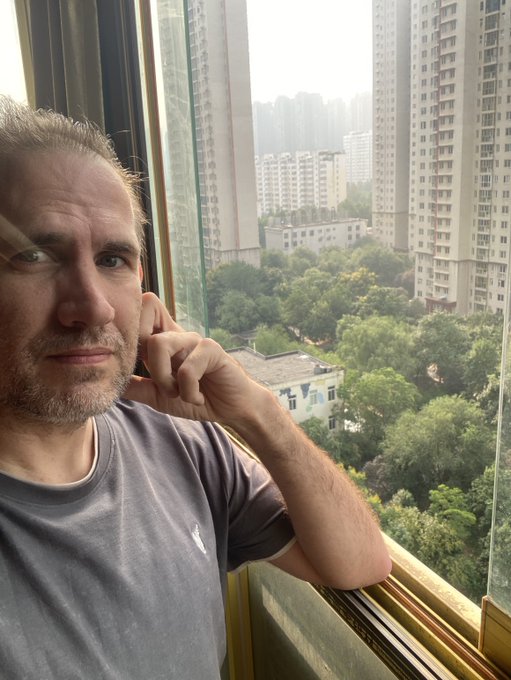 A man with short hair and a gray t-shirt leans against a window with his hand near his ear, looking out over a cityscape with high-rise buildings and trees.