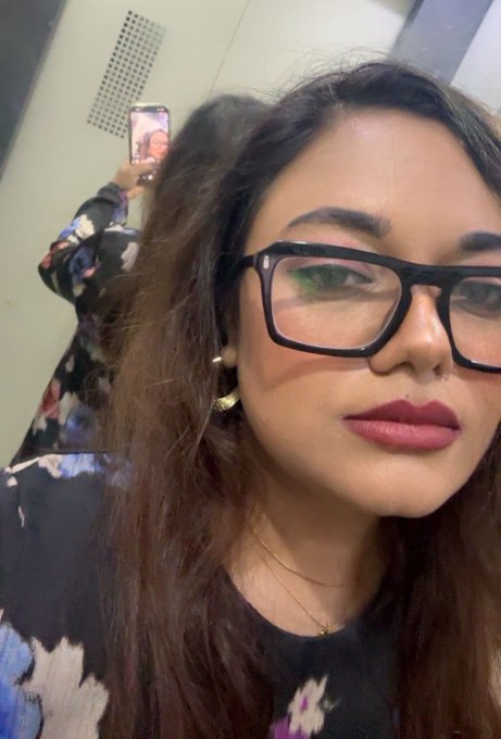 A selfie of a woman with long dark hair wearing black-rimmed glasses and a floral top, with a mirror reflection visible in the background.