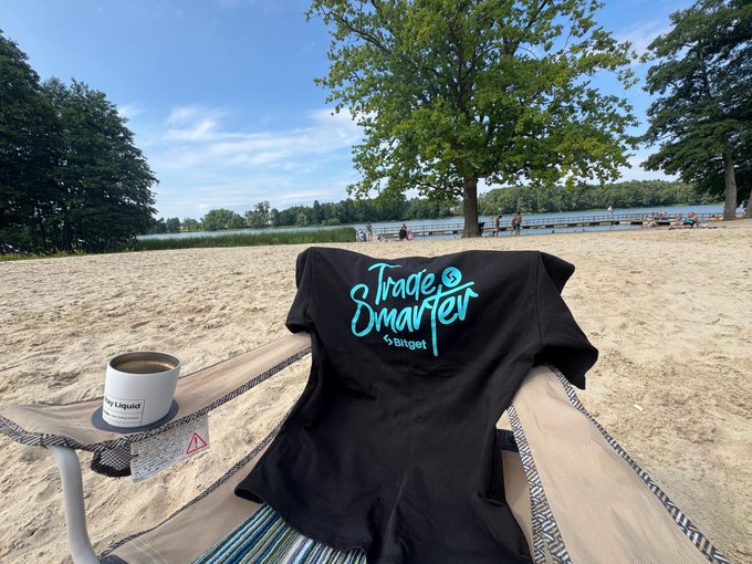 A black t-shirt reading 'Trade Smarter by BingX' draped over a beach chair next to a coffee mug labeled 'Liquid', overlooking a lake with people swimming.