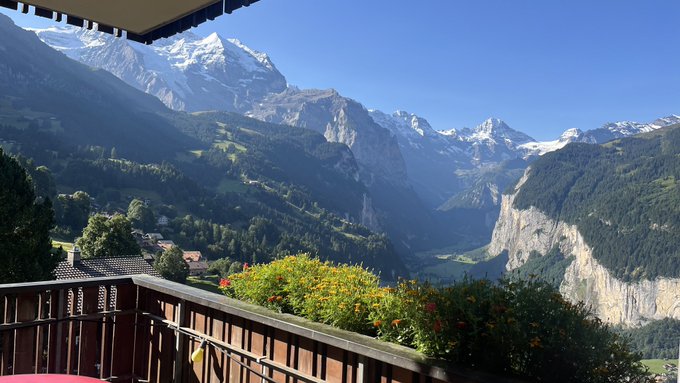 A scenic balcony view overlooks the snow-capped mountains and lush valleys of Wengen, Switzerland, with vibrant red flowers in the foreground. The serene alpine landscape evokes a peaceful Sunday morning, fitting the relaxed tone of the post before heading to church and engaging with BingX.