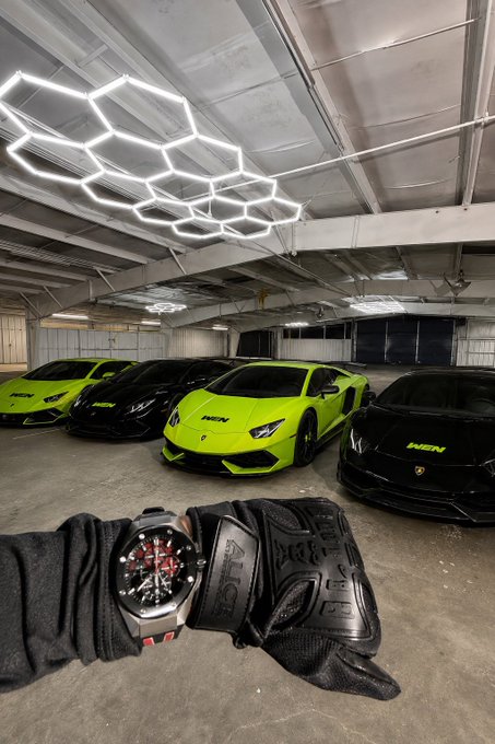 First-person view of a wrist wearing a luxury watch and black driving glove, with a row of four Lamborghini Huracáns (in green and black) parked in a garage under hexagonal LED ceiling lights.