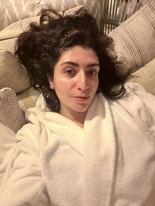 A selfie of a woman with curly dark hair wearing a white bathrobe, lounging against beige and patterned pillows.