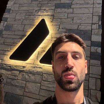 A selfie of a young man with a beard standing in front of an illuminated logo on a stone wall.