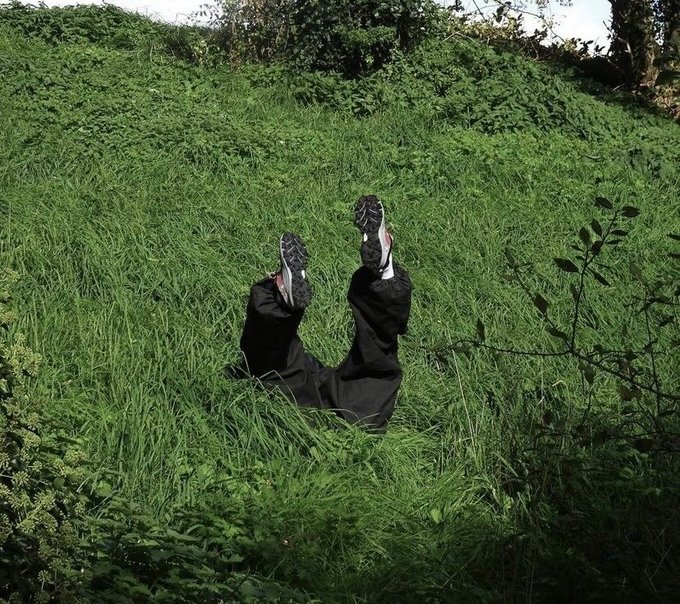 A person dressed in black lies upside down in tall green grass, with their legs and black-and-white sneakers sticking up in the air against a hillside.