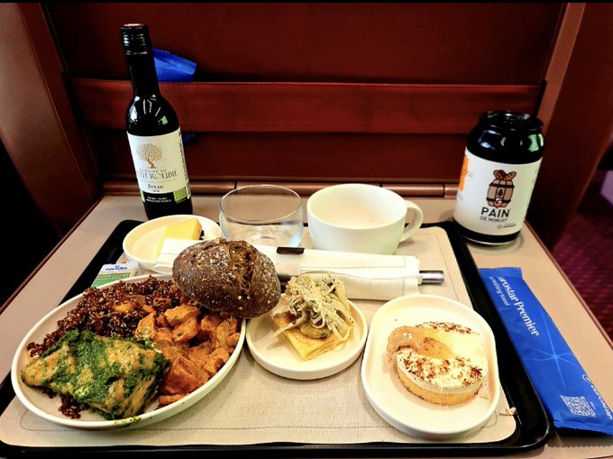 A first-class train meal across Europe features a tray with a bottle of wine, a craft beer can, an assorted plate of cold cuts and greens, a small mushroom roll, cheese, and a cinnamon roll, accompanied by a blue "Premium/Privilège" sleeve. The spread complements the post's celebration of a quiet, scenic European rail journey, setting the stage for enjoying a drink while diving into the charts.