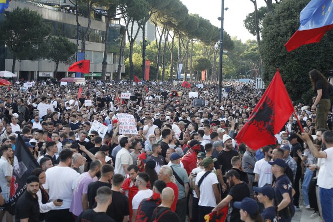 iced tea and soda stands line a tree-lined street, with protesters holding red flags and banners during a protest march.