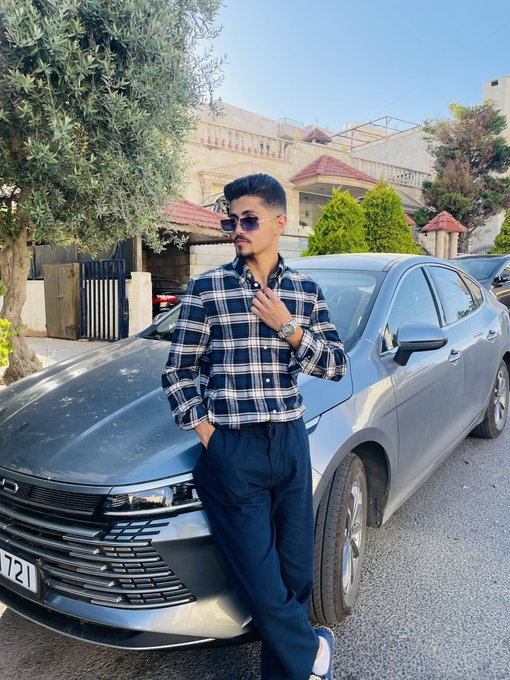 A man in a navy plaid shirt and sunglasses leans against a gray sedan parked on a residential street.