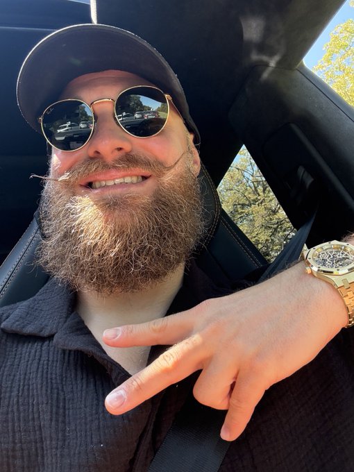 A bearded man wearing a cap and round sunglasses takes a selfie inside a car, flashing a peace sign while showing off a gold watch on his wrist—chilling and unbothered as he greets the crypto market with "GM to everyone getting ready for $30k lows."