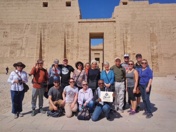 A group of tourists poses in front of the ancient Egyptian Medinet Habu temple, with one person holding a sign reading "TANSIQ." The image appears to be used ironically or in juxtaposition with the post, as the post discusses concerns about the TANSIQ project possibly being a scam, while the photo shows a completely unrelated group sightseeing at a historic Egyptian landmark rather than any mining or project-related activity.