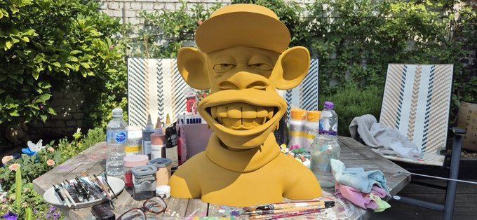 A yellow sculpted bust of a cartoon character with an exaggerated grin and prominent teeth, sitting on a table surrounded by paint bottles and art supplies in an outdoor garden setting.