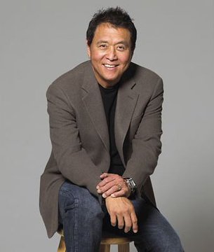 A portrait photo of an Asian man smiling, wearing a brown blazer over a black shirt and jeans, seated on a stool