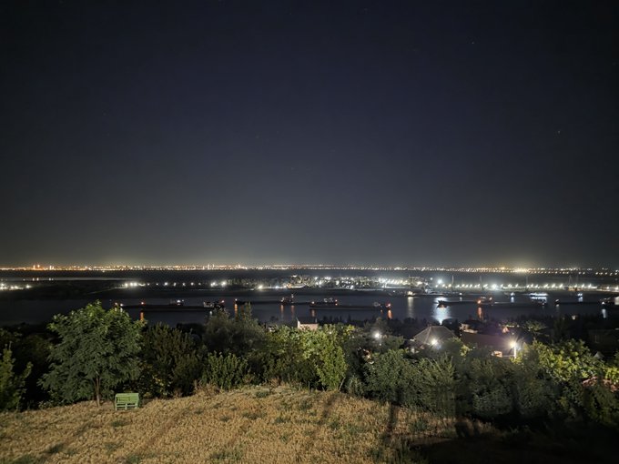 A nighttime view from an elevated vantage point shows city lights twinkling across a body of water, with a dark grassy field and a small bench in the foreground.