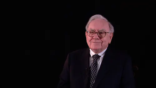 An elderly man with white hair and glasses, wearing a dark suit and patterned tie, is shown against a black background.
