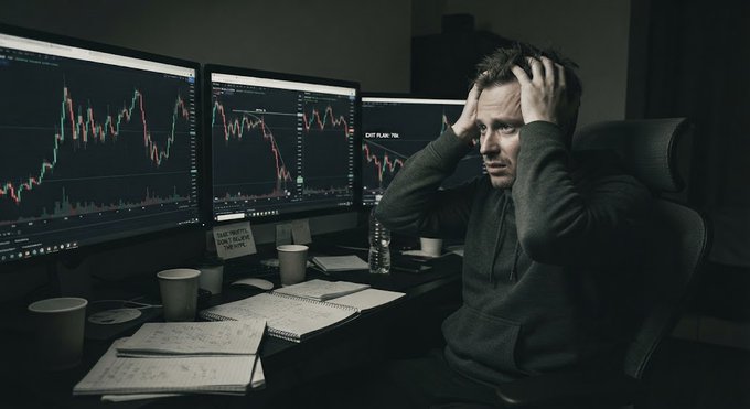 A man sitting at a desk in a dark room, holding his head in apparent distress while looking at three monitors displaying stock market charts.