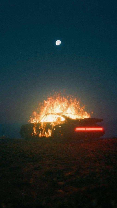 A car engulfed in flames at night under a dark sky with a visible full moon, casting an orange glow on the surrounding ground.