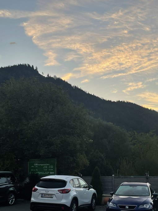 A scenic view at dusk showing forested mountains under a cloudy sky, with parked cars including a white Mazda in the foreground and a green 'Öffnungszeiten' information sign.