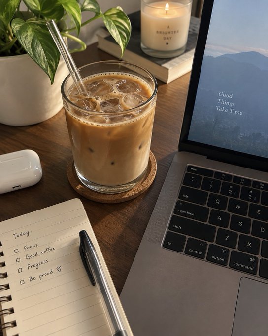 A cozy desk setup featuring an iced coffee, a lit candle, a plant, a notebook with a to-do list ("Today: Focus, Good coffee, Progress, Be proud"), and a laptop displaying "Good Things Take Time" on its screen.