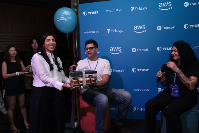 A woman in a white shirt holds a clear box of baked goods at an event with a backdrop featuring logos for Trust, Mercuryo, Yield.xyz, AWS, and Transak.
