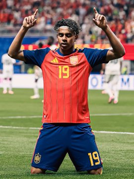 A soccer player wearing the red Spain national team jersey with number 19 kneeling on the field and pointing both index fingers upward in celebration.
