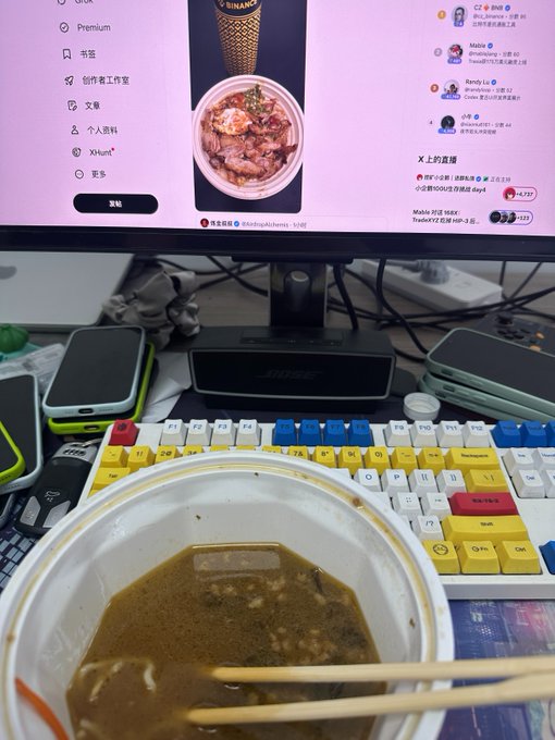 A desk setup showing a bowl of dark broth/sauce with chopsticks in front of a colorful mechanical keyboard, with a computer monitor displaying a live stream featuring food content and chat.