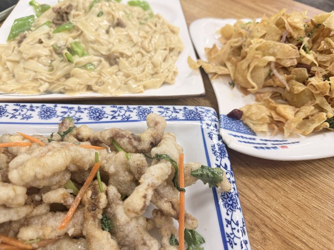 Three Chinese dishes served on blue and white patterned plates, including stir-fried noodles with vegetables, crispy fried noodles, and a battered fried dish garnished with carrots and herbs.