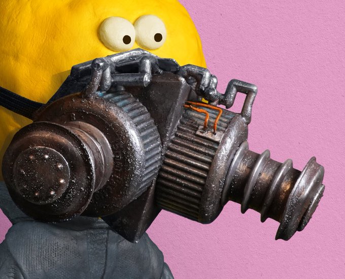 A photo of a yellow Minion plush toy wearing a detailed metallic steampunk-style respirator mask against a pink wall.