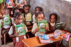 A group of young school children in uniforms sit at wooden desks, each holding up a colorful school workbook or textbook in a classroom.