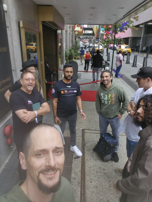 Group selfie photo taken outside a building entrance, featuring several people including a man in a black '90' commemorative t-shirt, with a red carpet and yellow taxi visible on the street behind them.