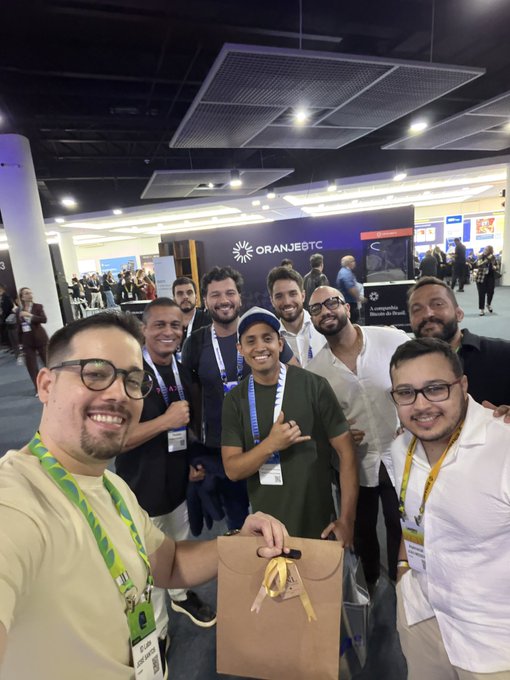 A group selfie of seven smiling people at a convention center, with an 'ORANGEBTC' booth visible in the background and a brown gift bag held in the center.