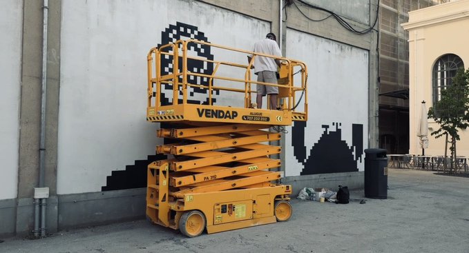 A worker on a yellow VENDAP scissor lift is creating a black pixel-art style mural on a large white building wall, with another building featuring arched windows visible in the background.