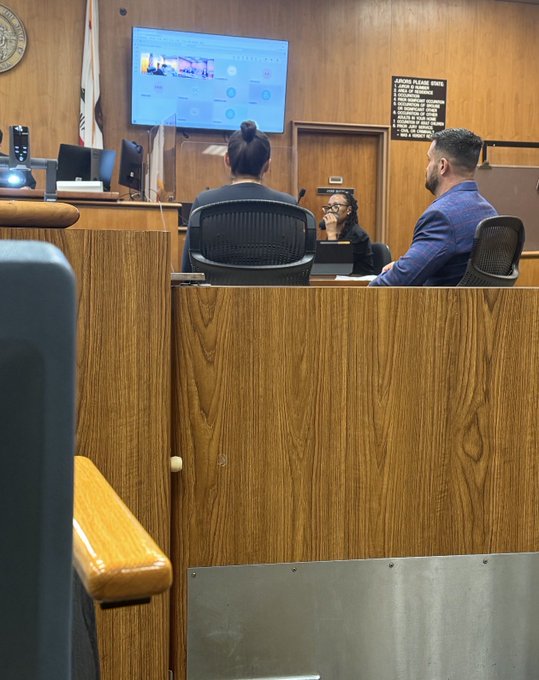 A courtroom scene viewed from behind wooden barriers, showing a man in a blue suit and a woman with glasses seated at a table, with a large screen displaying images mounted on the wood-paneled wall, and what appears to be a bailiff or clerk at the bench.