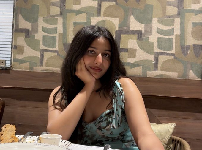 A young woman with long dark hair, wearing a floral green dress, rests her chin on her hand at a restaurant table, appearing thoughtful and approachable. The caption playfully references her pensive, welcoming expression, suggesting she's hoping to meet a patient mentor willing to teach her the risks of leverage trading.