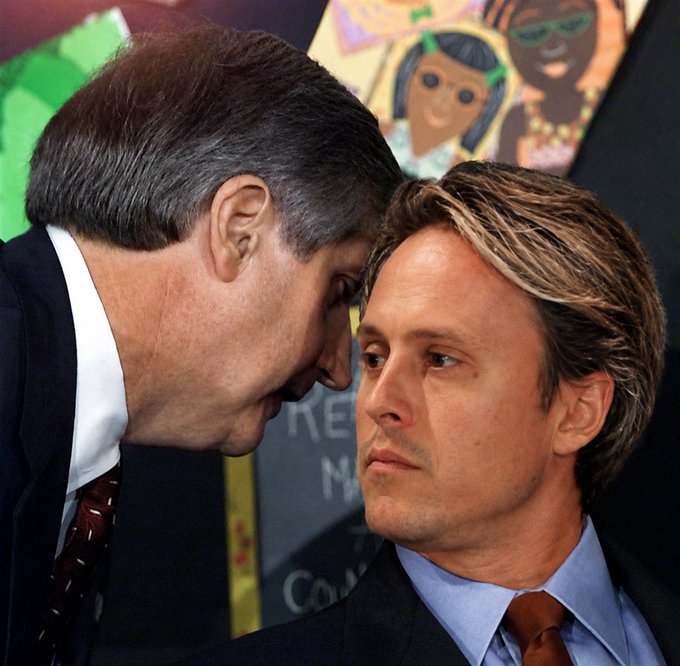 A photograph of two men in suits standing close together in profile view, with children's artwork visible in the background.