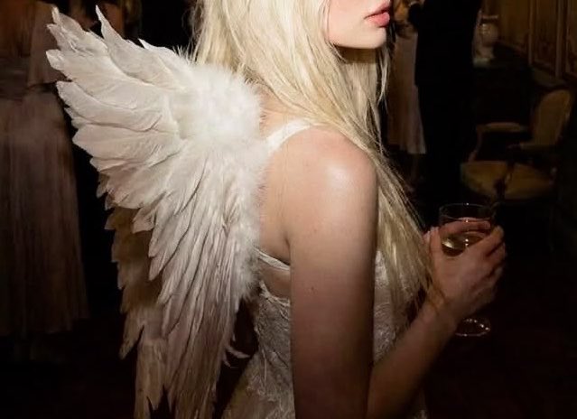 A woman with long blonde hair wears white angel wings and a white dress while holding a wine glass in an elegant interior setting.