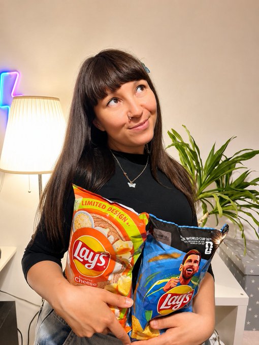 A woman with dark hair and bangs smiles while holding two bags of Lay's potato chips, including a 'Cheese' flavor and a FIFA World Cup 26 limited edition bag featuring a player.