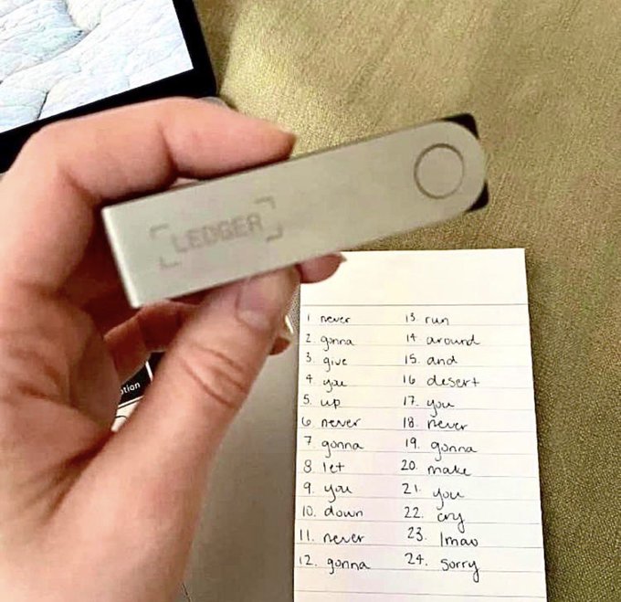 A hand holds a silver Ledger hardware wallet above a handwritten numbered list of 24 words/phrases (including "never," "gonna," "give," "you," "up"), which appears to be a cryptocurrency seed recovery phrase backup.