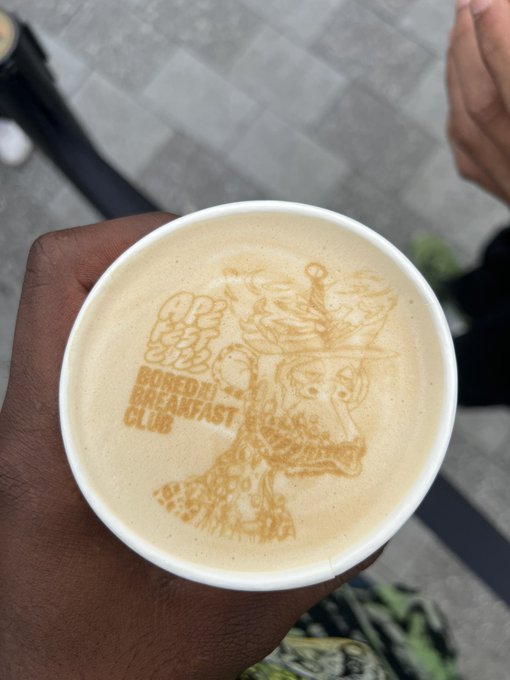 A close-up of a latte held by a dark-skinned hand, featuring intricate foam art of a crowned character alongside the logo 'BORED APE BREAKFAST CLUB'.