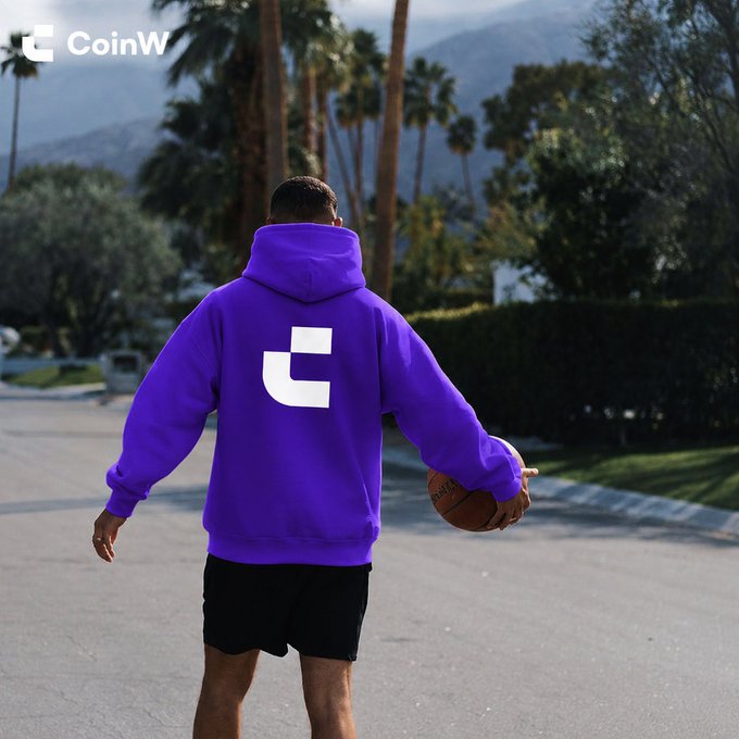 A person seen from behind wearing a purple hoodie featuring a white logo, holding a basketball while walking down a palm tree-lined street with the 'CoinW' logo in the top left corner.