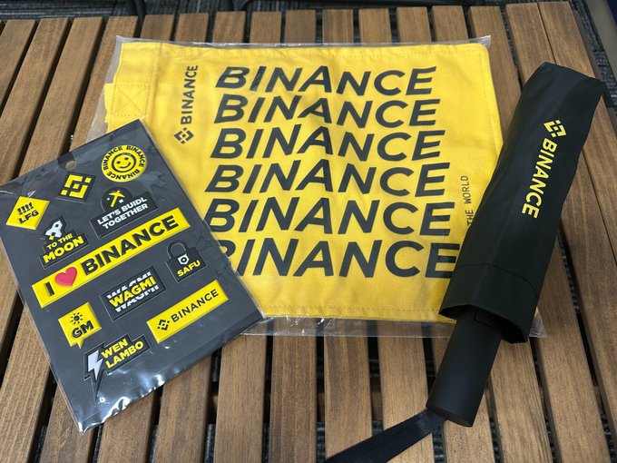 The image features a black umbrella with the word "binance" written on it, along with some stickers attached to it. The umbrella has a handle at one end for easy carrying. There are also several stickers placed around the umbrella, including one that says "I love binance". The overall scene suggests that someone might be using these items as part of their daily routine or as a way to express their support and appreciation for the company.