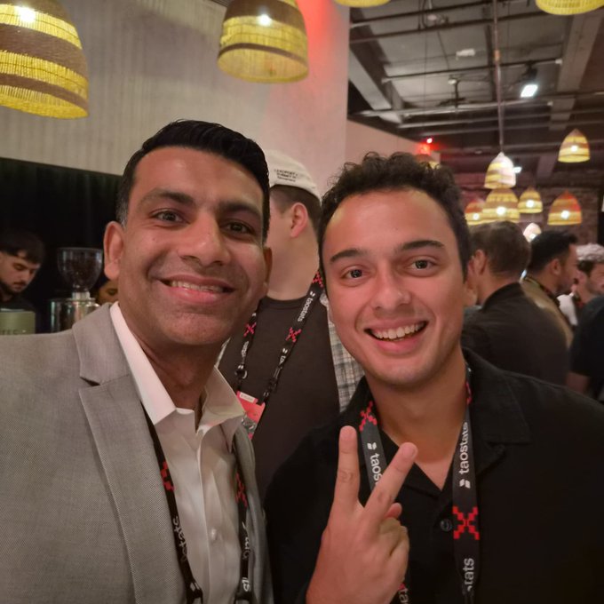 Two smiling men wearing 'fatbatter' lanyards pose for a selfie at an indoor event, with one holding up a peace sign amid a crowd and decorative hanging lamps.