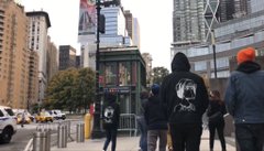 A street scene in what appears to be a city (likely New York near Central Park based on the setting), showing pedestrians walking on a sidewalk with a person in a black hoodie featuring a white circular logo on the back, with tall buildings, traffic, and a green kiosk visible in the background.