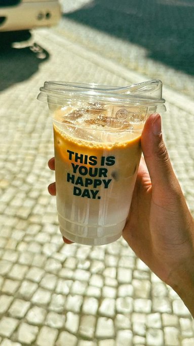 A hand holds a clear plastic cup of iced coffee with the text "THIS IS YOUR HAPPY DAY." printed on it, photographed on a sunlit cobblestone street.