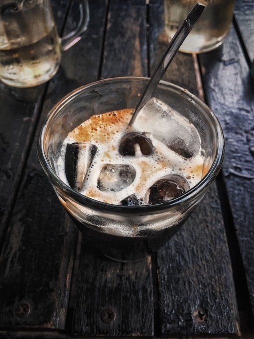 A glass of iced coffee or iced tea with large ice cubes and a black straw sits on a dark, weathered wooden table.