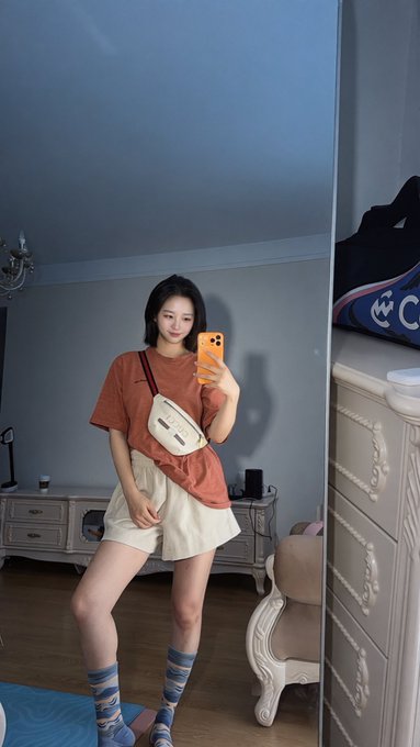 A mirror selfie of a young Asian woman in a bedroom wearing an oversized rust-colored t-shirt, cream shorts, and blue patterned knee socks. She carries a cream crossbody bag and holds an orange phone to take the photo, with a dresser and bed visible in the room.