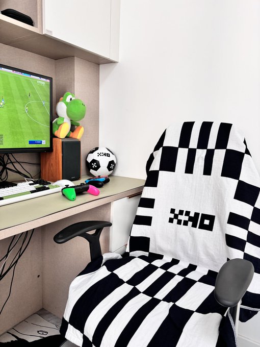 Photo of a minimalist computer gaming/setup corner featuring a Yoshi plush toy, a soccer ball, gaming controllers on a desk with a monitor displaying a soccer game, and a black and white checkered blanket with "XCHO" text draped over it.